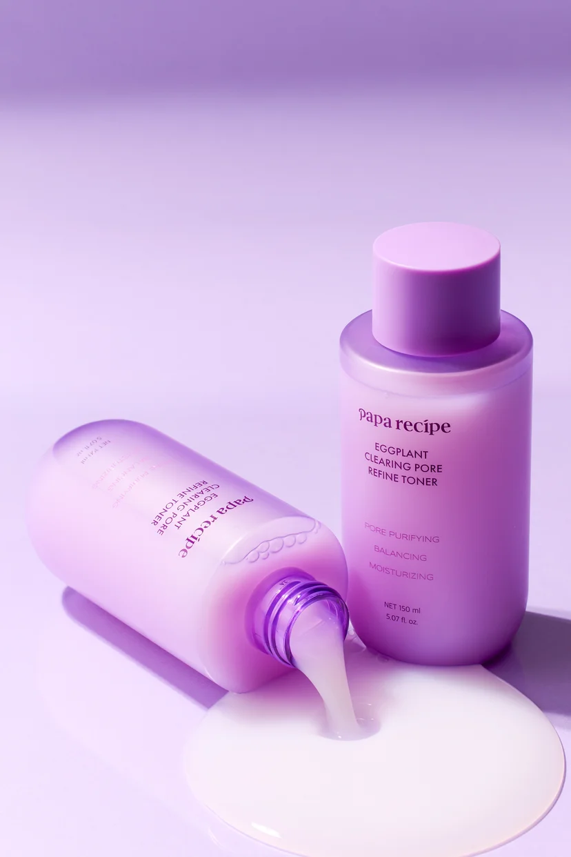 Two Papa Recipe Eggplant Clearing Pore Refine Toner 150ml bottles on a lavender surface — one standing upright and one tipped on its side with the cap open, pouring a pool of milky white toner onto the surface