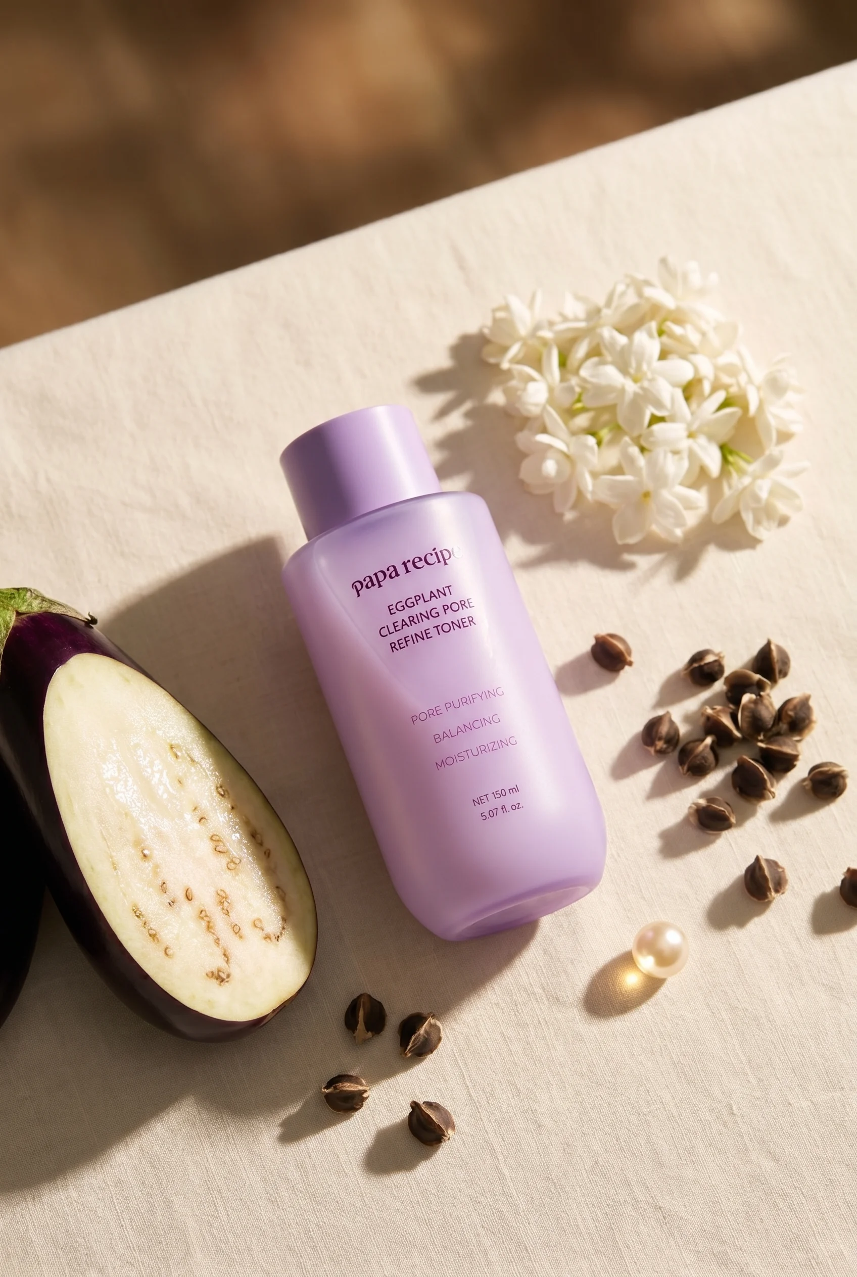 Papa Recipe Eggplant Clearing Pore Refine Toner 150ml bottle laid flat on a linen surface, surrounded by a halved eggplant, white flowers, small dried seeds, and a pearl