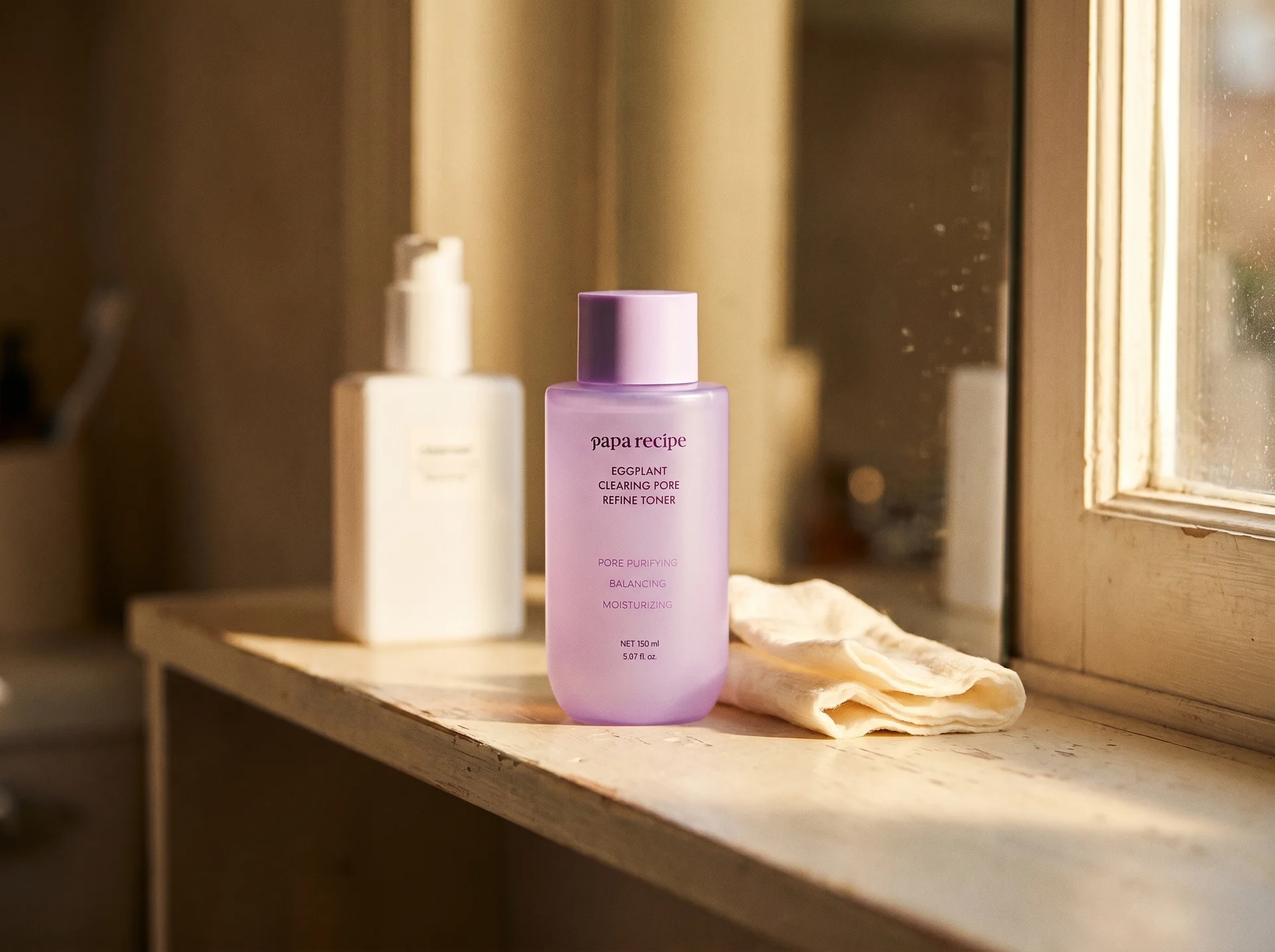 Papa Recipe Eggplant Clearing Pore Refine Toner 150ml bottle placed on a worn wooden shelf beside a white skincare bottle and a folded linen cloth, lit by warm natural light from a nearby window