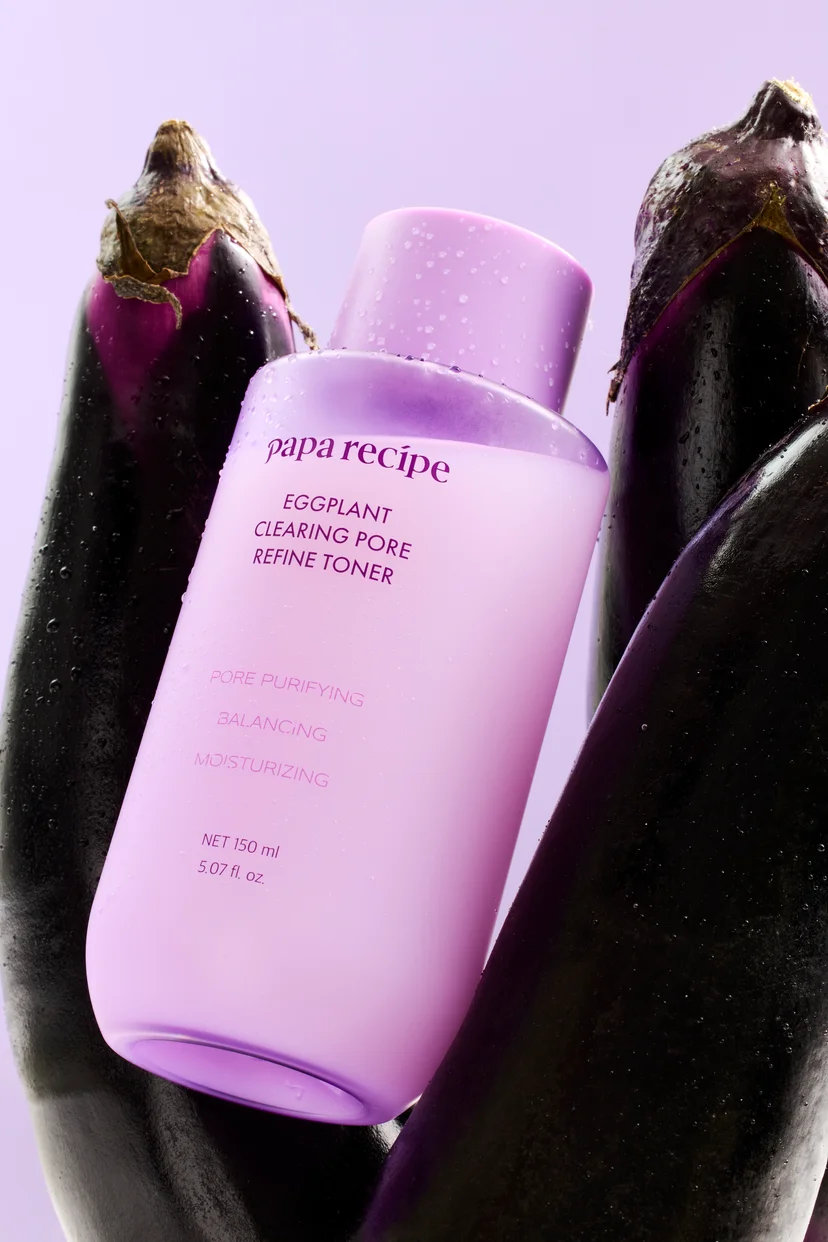 Papa Recipe Eggplant Clearing Pore Refine Toner 150ml bottle with water droplets, nestled between two whole fresh eggplants against a lavender background