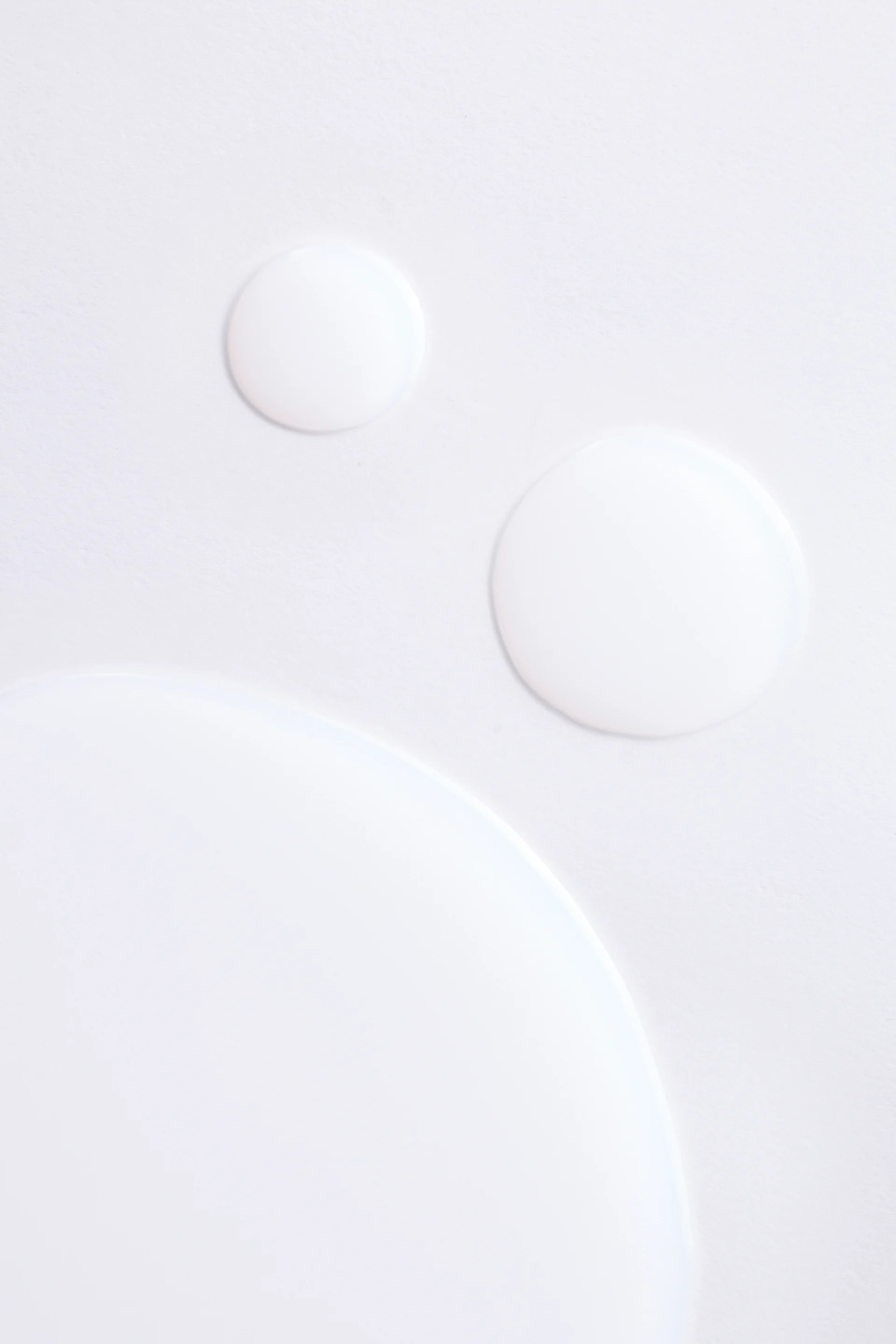 Close-up of multiple milky white toner drops in varying sizes pooled on a white surface, showing the lightweight texture of the formula