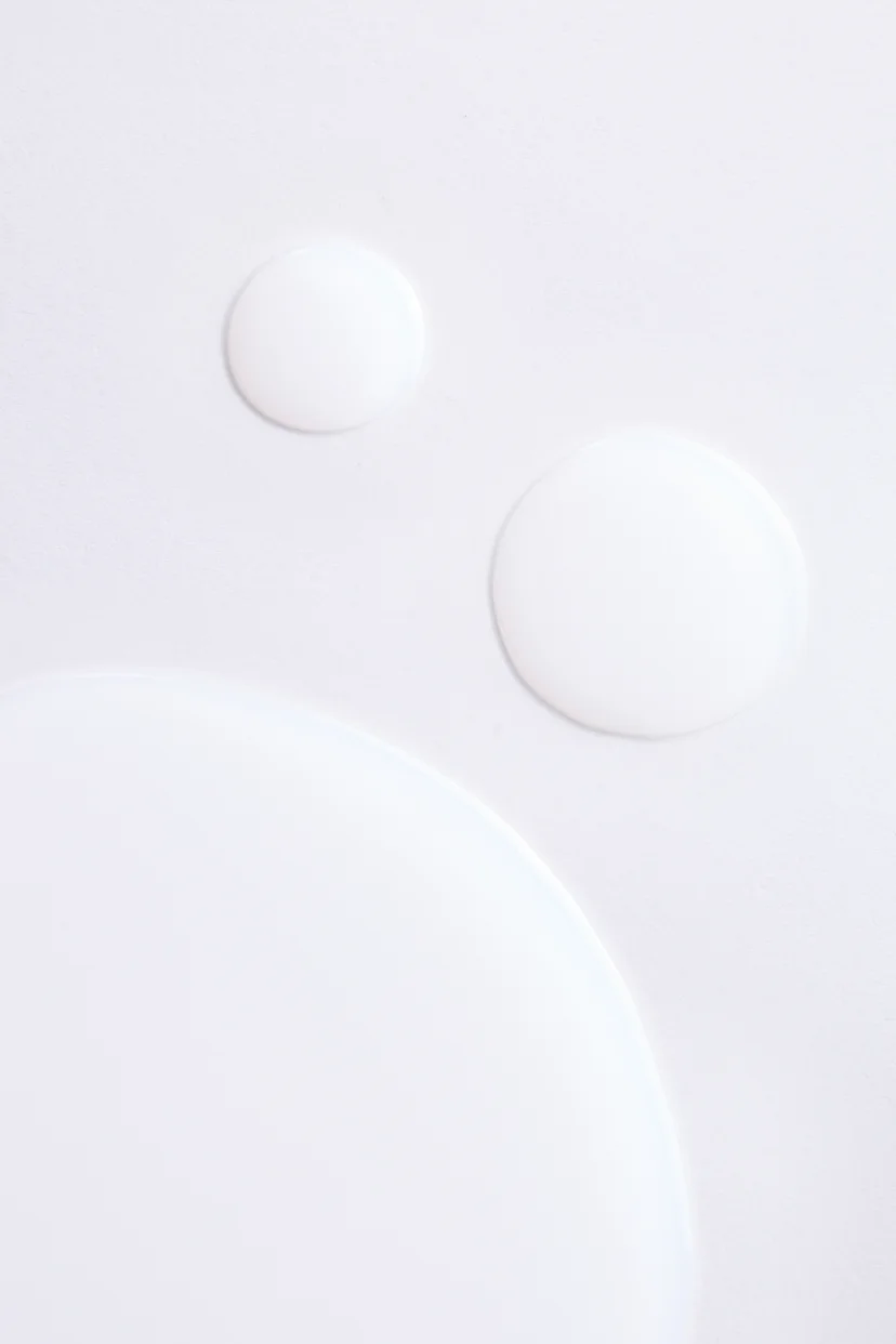 Close-up of multiple milky white toner drops in varying sizes pooled on a white surface, showing the lightweight texture of the formula