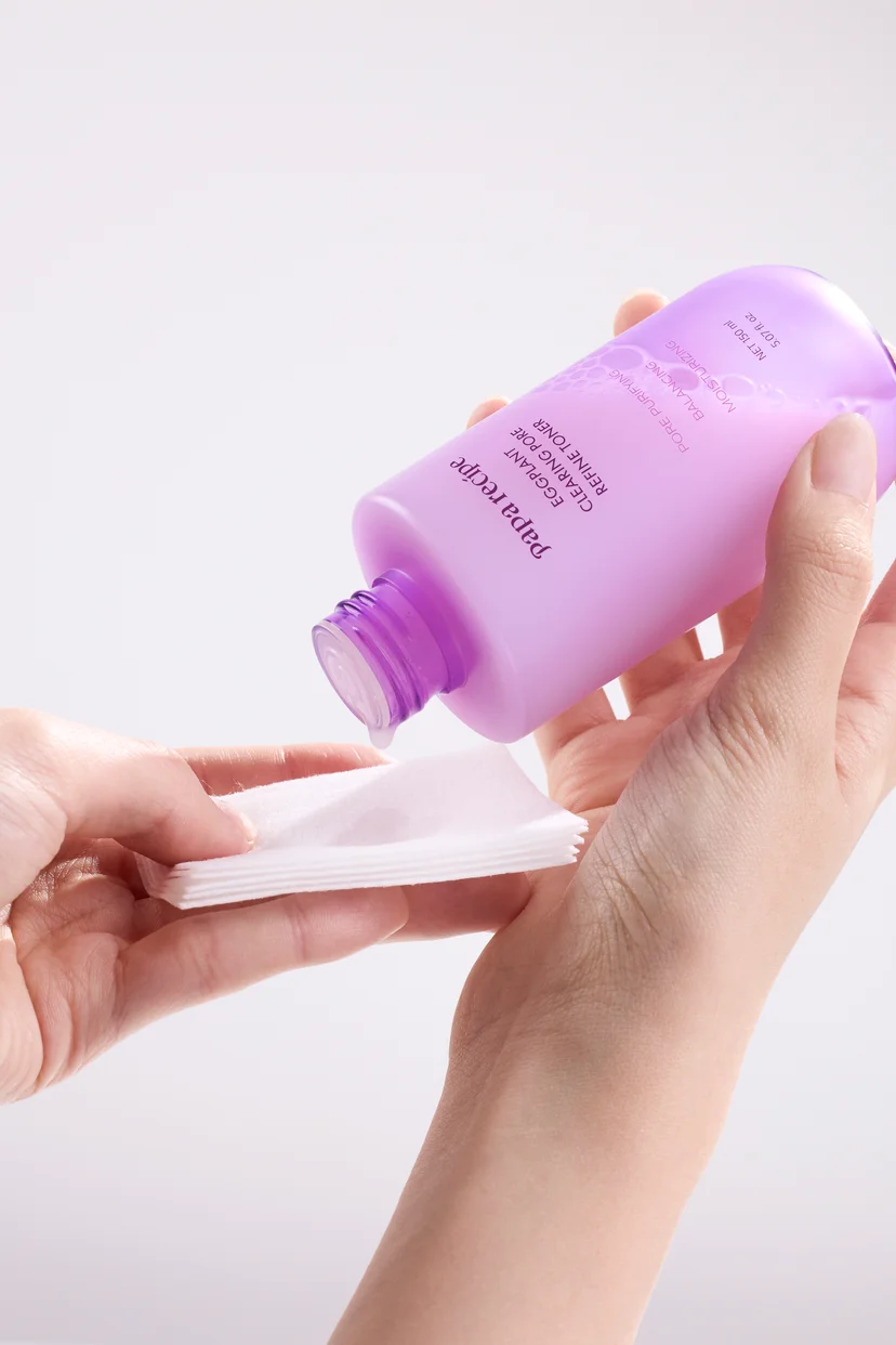 Two hands demonstrating how to apply Papa Recipe Eggplant Clearing Pore Refine Toner — one hand holding a cotton pad while the other pours the milky toner directly from the upside-down bottle onto the pad, against a white background