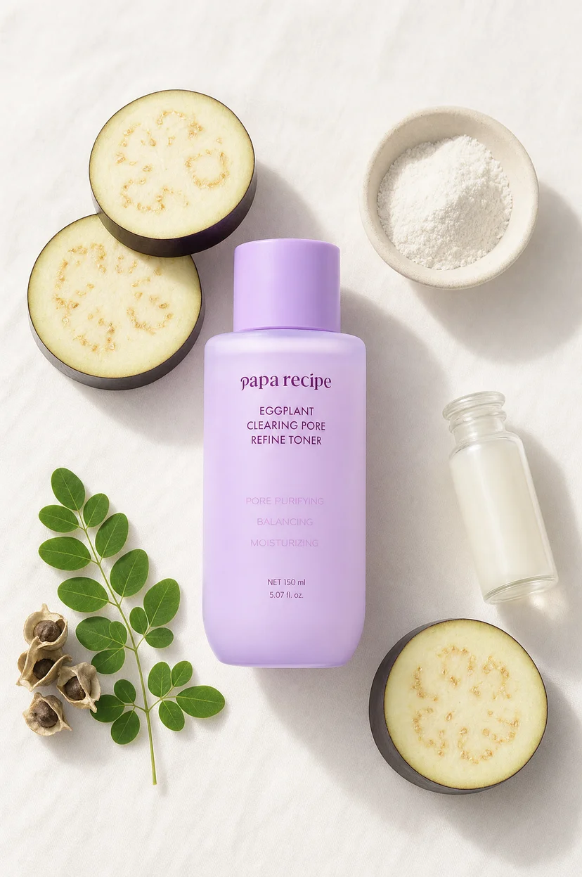 Papa Recipe Eggplant Clearing Pore Refine Toner 150ml bottle on a white linen surface, surrounded by sliced eggplant, a bowl of white powder, a small glass bottle with milky liquid, green leaves, and dried seeds