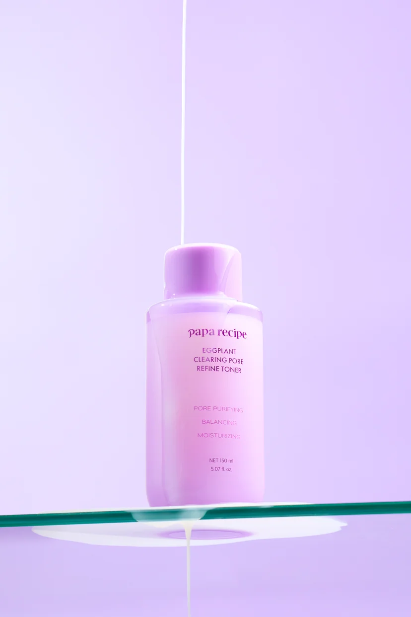 Papa Recipe Eggplant Clearing Pore Refine Toner 150ml bottle on a glass shelf against a lavender background, with a thin stream of milky white toner pouring down from above onto the bottle and dripping off the shelf below
