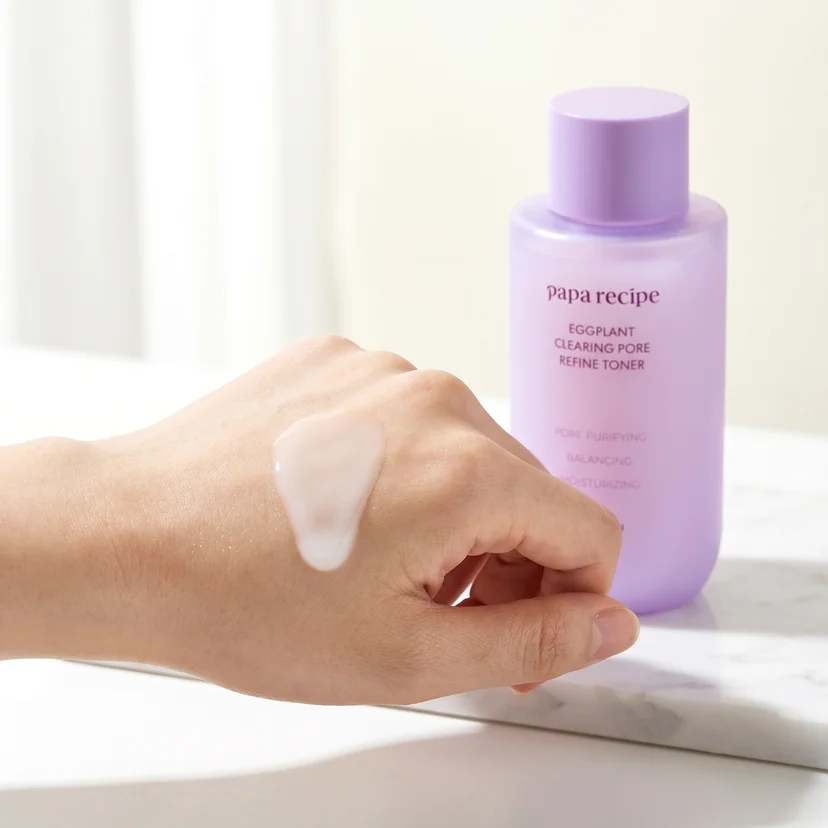 A milky white toner swatch spread across the back of a hand, with a Papa Recipe Eggplant Clearing Pore Refine Toner 150ml bottle softly blurred in the background on a marble surface