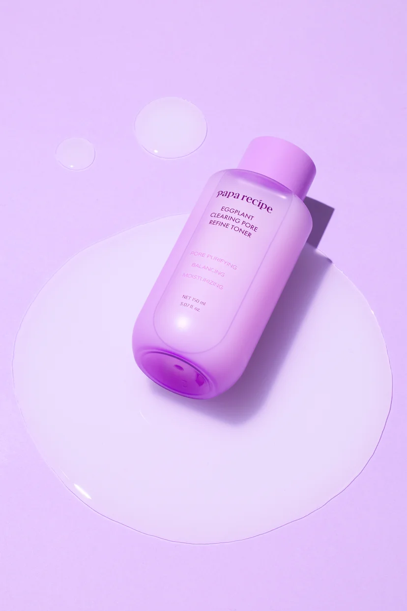 Papa Recipe Eggplant Clearing Pore Refine Toner 150ml bottle lying on its side atop a large circular milky toner puddle, with two smaller toner droplets scattered on a lavender background