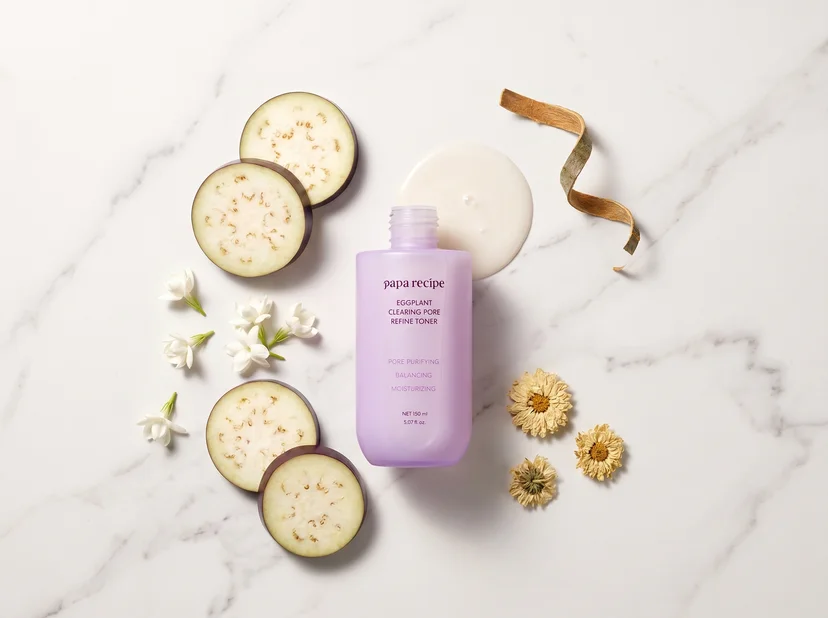 Papa Recipe Eggplant Clearing Pore Refine Toner 150ml bottle without cap, surrounded by sliced eggplant rounds, white jasmine flowers, dried chrysanthemum, and a curled strip of willow bark, with a milky white toner pool behind the bottle on a marble surface