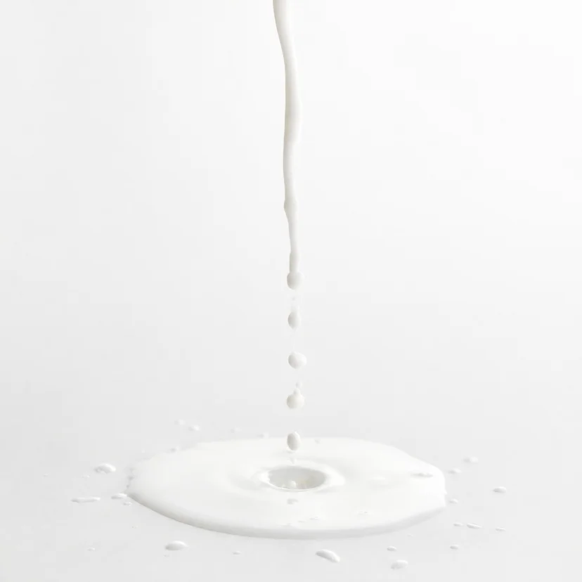 A thin stream of milky white toner pouring downward into a pooling splash on a white surface, with small droplets scattered around the edges