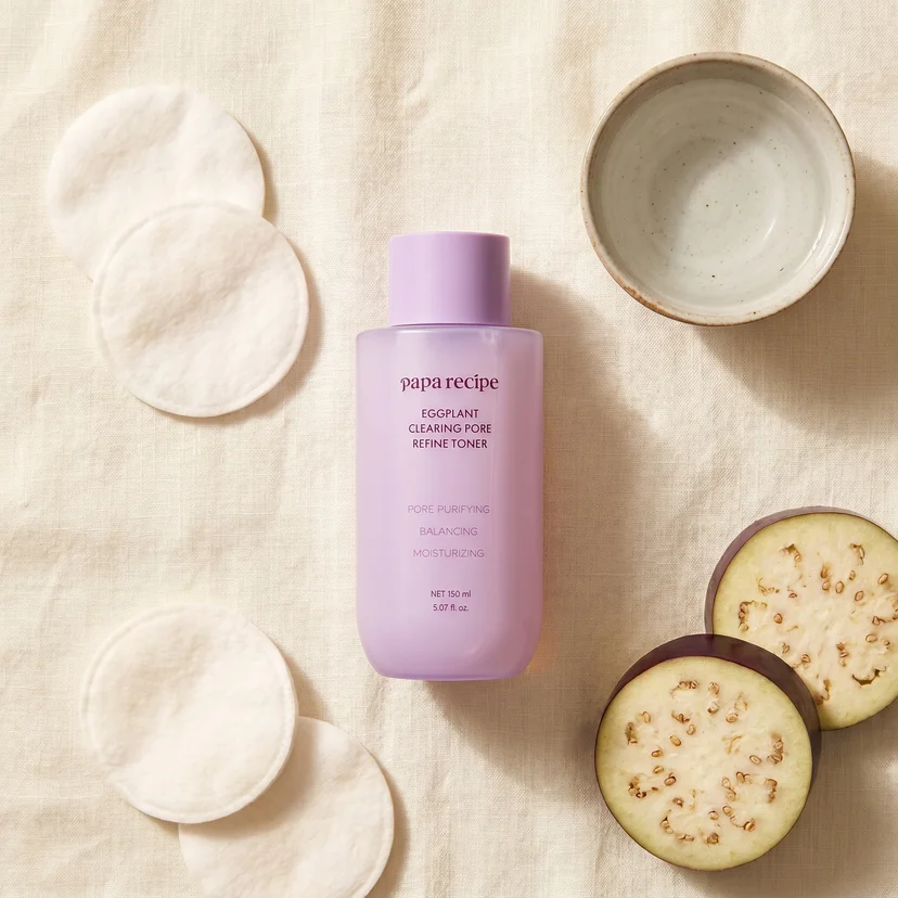 Papa Recipe Eggplant Clearing Pore Refine Toner 150ml bottle centered on a linen surface, flanked by three round cotton pads on the left, sliced eggplant rounds on the lower right, and a small ceramic bowl on the upper right