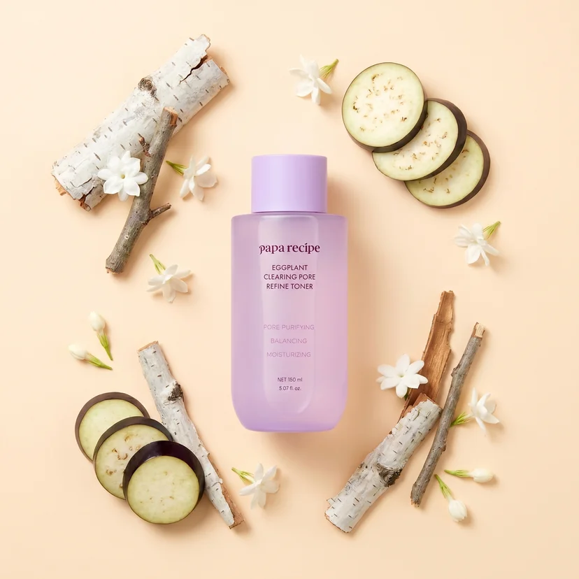Papa Recipe Eggplant Clearing Pore Refine Toner 150ml bottle centered on a peach background, encircled by sliced eggplant rounds, white birch bark sticks, and white jasmine flowers arranged in a ring pattern