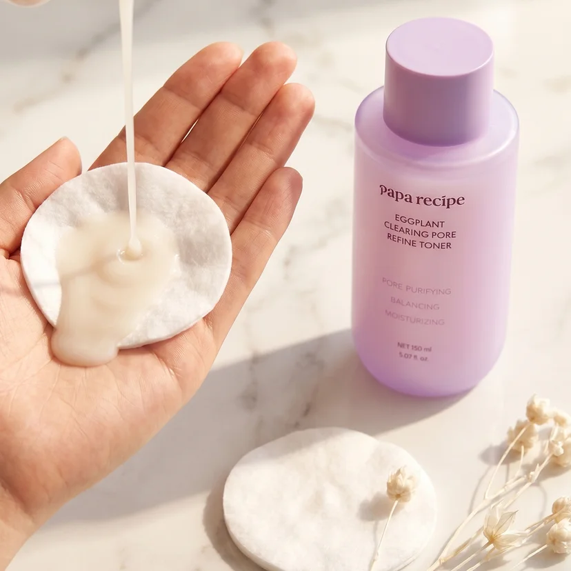 A hand holding a round cotton pad while milky white toner is poured directly onto it from above, with a Papa Recipe Eggplant Clearing Pore Refine Toner 150ml bottle and dried white flowers on a marble surface in the background