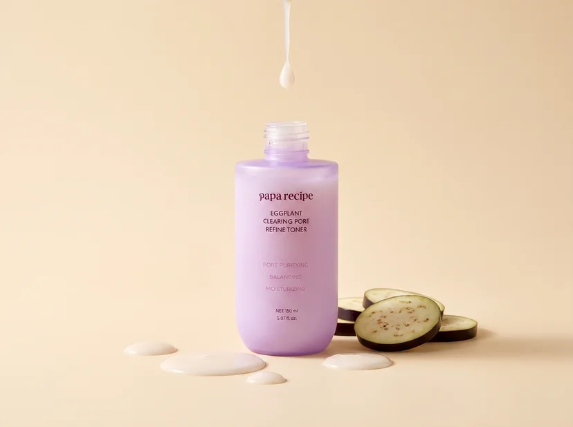 Papa Recipe Eggplant Clearing Pore Refine Toner 150ml open bottle without cap, with a single drop of milky white toner falling from above into the bottle opening, milky toner droplets pooled in front, and sliced eggplant rounds beside the bottle on a peach background