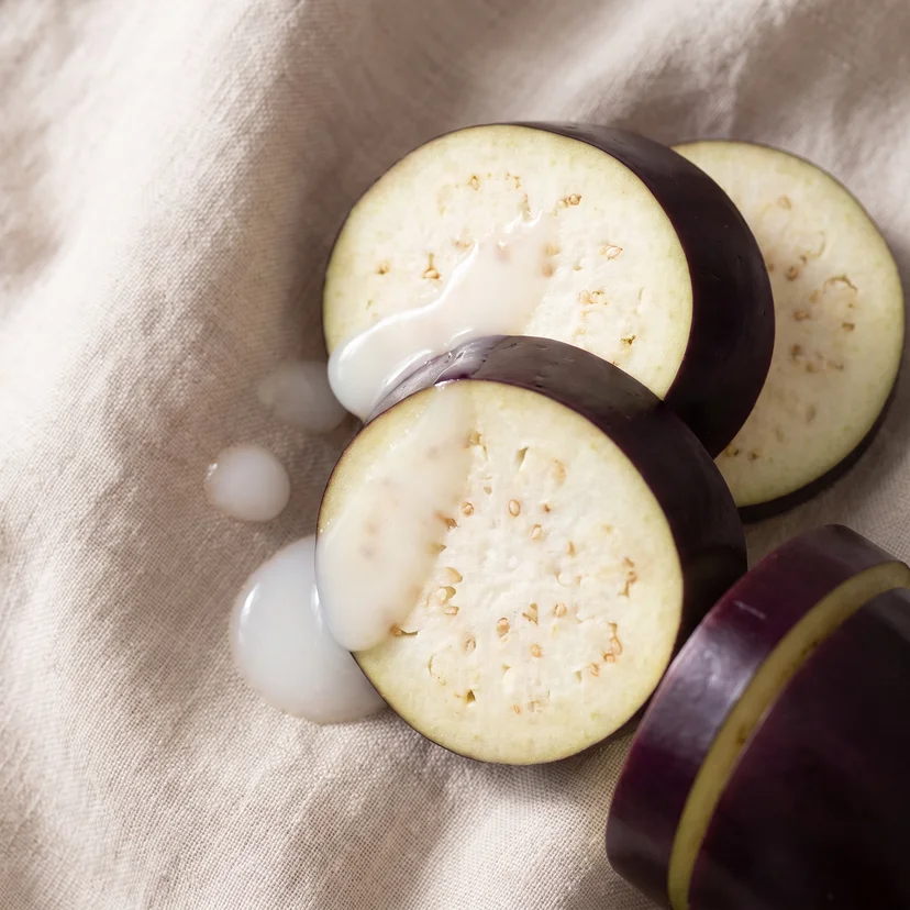 Close-up of three cross-sectioned eggplant rounds stacked on a linen surface, with milky white toner poured over them and dripping down the sides, and a whole eggplant slice in the lower right corner