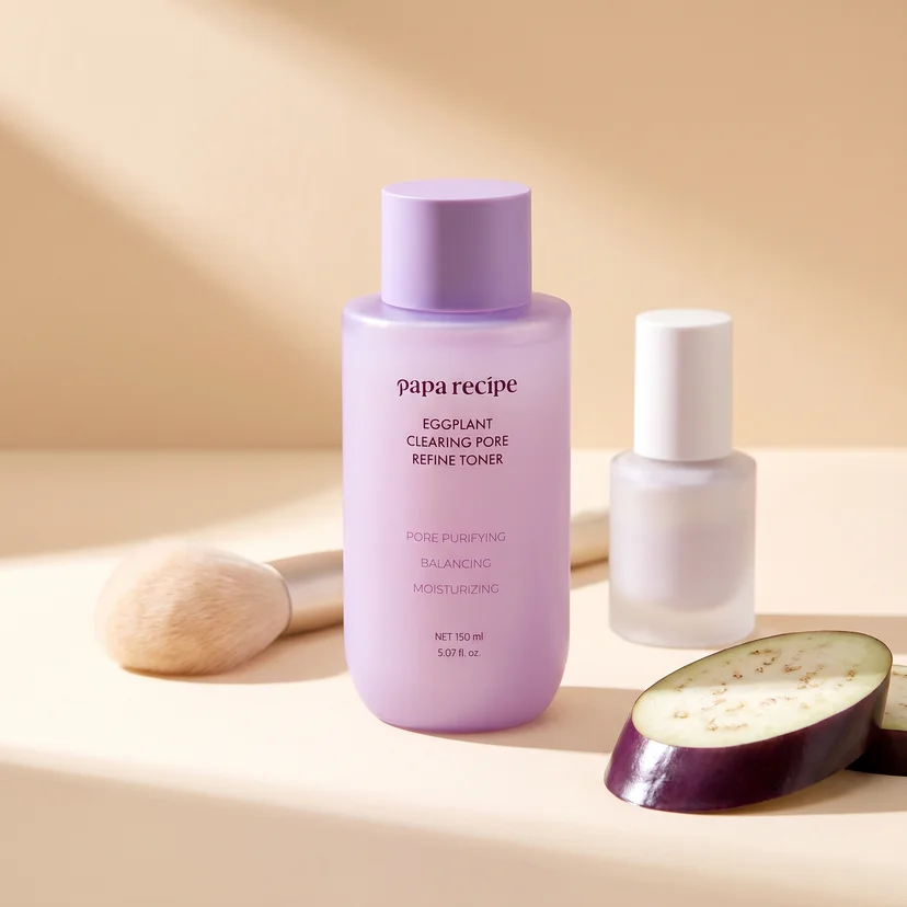 Papa Recipe Eggplant Clearing Pore Refine Toner 150ml bottle centered on a warm beige surface, flanked by a makeup brush on the left, a frosted glass primer bottle on the right, and a sliced eggplant in the foreground