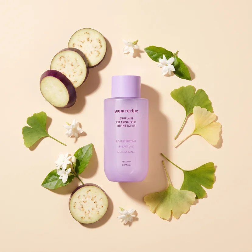 Papa Recipe Eggplant Clearing Pore Refine Toner 150ml bottle centered on a peach background, encircled by sliced eggplant rounds, white jasmine flowers with leaves, and multiple ginkgo biloba leaves arranged in a ring