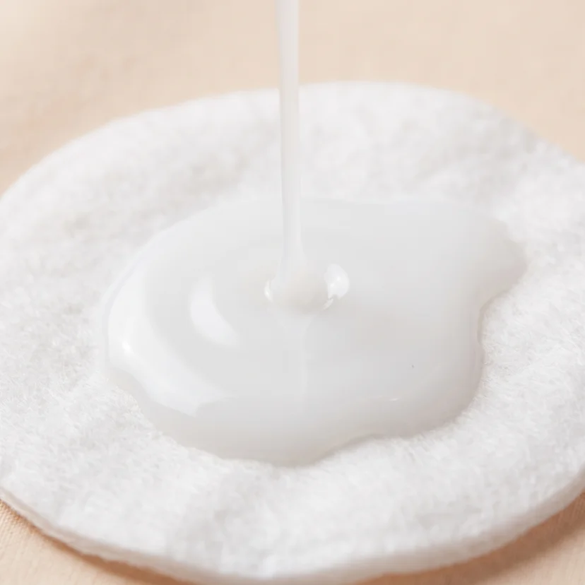 Close-up of milky white toner being poured onto a soft cotton round, showing its smooth, lightly opaque consistency.