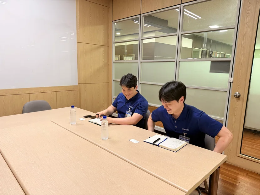 유니크(UNICK) 로고가 새겨진 남색 폴로셔츠를 입은 두 담당자가 회의실 테이블에 앉아 인터뷰하고 있다. 왼쪽이 박용한 매니저, 오른쪽이 박찬건 선임매니저.