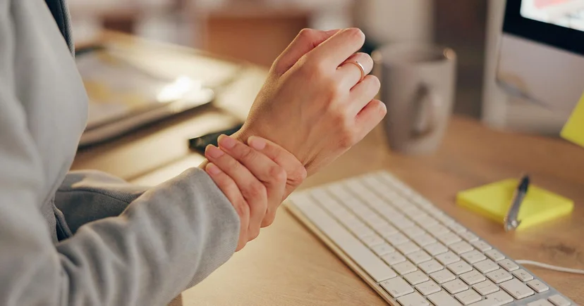 잦은 키보드 타이핑과 마우스 사용으로 인해 엄지손가락 쪽 손목에 찌릿한 통증을 느끼는 직장인의 모습