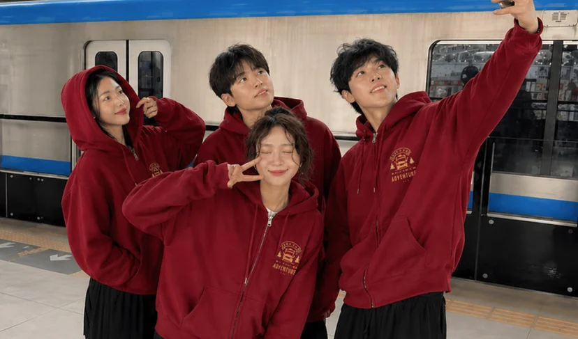 마플 젤란 라이트 후드집업 – 여행 크루 로고를 가슴에 인쇄한 버건디 후드집업을 남녀 4명이 맞춰 입고 지하철 승강장에서 셀카를 촬영하는 이미지