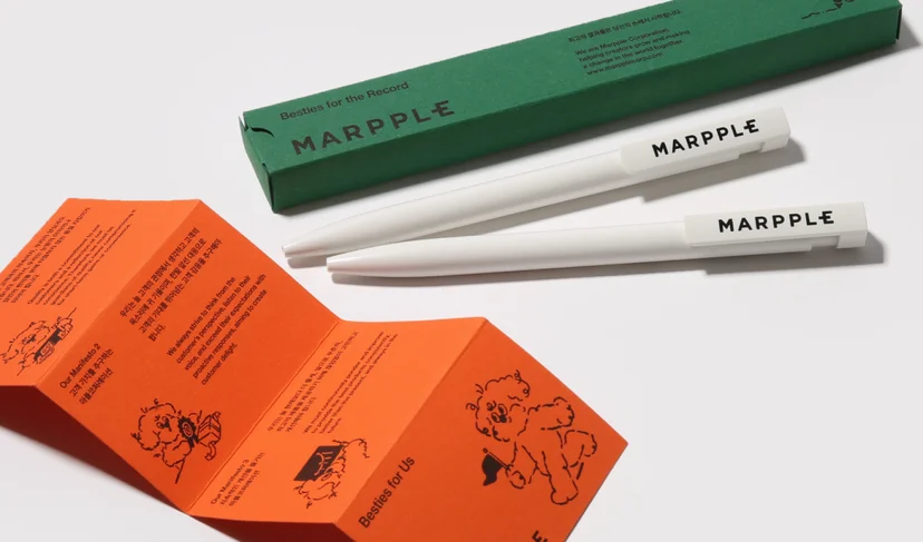 마플 사내 굿즈 – MARPPLE 로고를 인쇄한 흰색 볼펜 2개와 초록색 전용 패키지, 브랜드 소개가 적힌 주황색 접지 카드를 흰 배경에 배치한 이미지