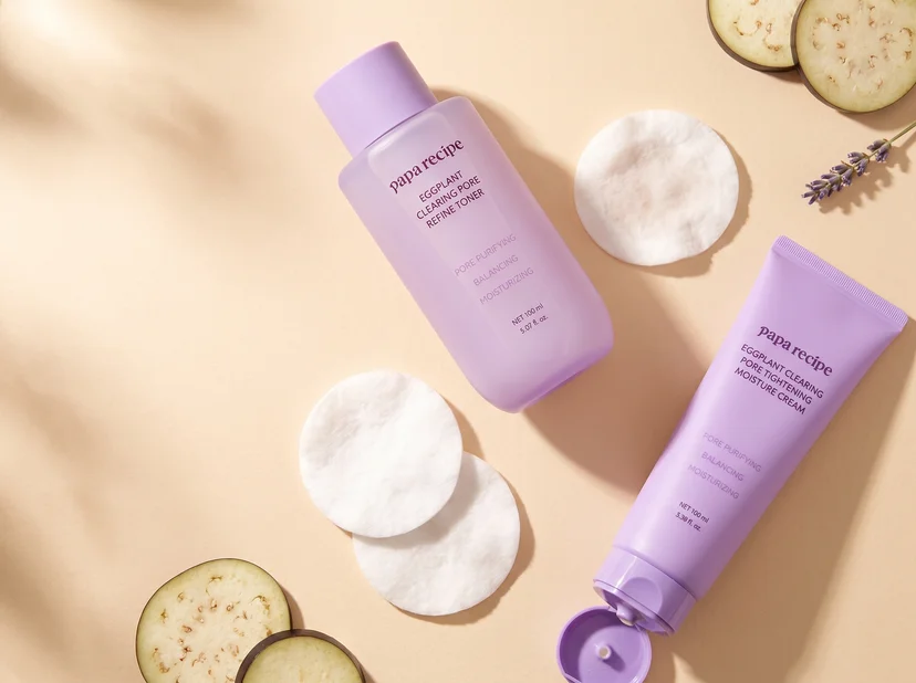 Flatlay arrangement of Papa Recipe pore refine milky toner, pore tightening moisture cream, cotton pads, eggplant slices, and lavender sprigs on a neutral surface.