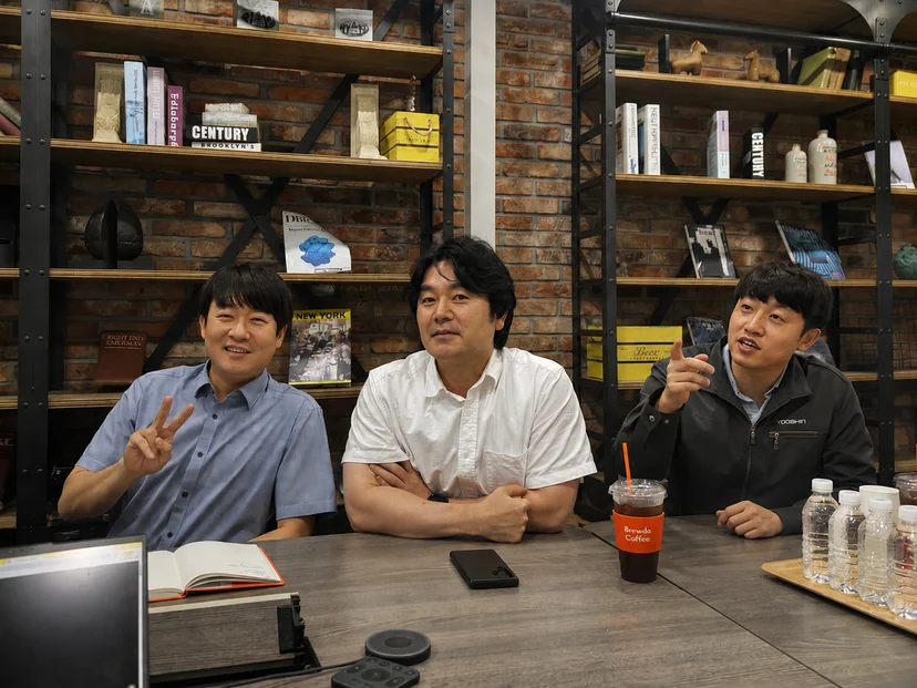 유신 재무팀 임직원 세 명이 카페형 회의 공간의 테이블에 나란히 앉아 인터뷰에 응하는 모습. 벽돌 벽과 책이 놓인 선반이 배경이고, 테이블에는 음료와 녹음기가 놓여 있다.