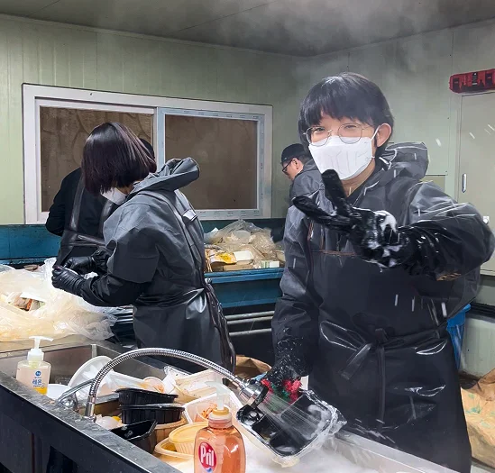 싱크대 수조에서 검은색 방수복을 입고 세제 거품으로 플라스틱 용기를 씻으며 브이 포즈를 취하는 작업자
