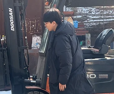 검은색 롱패딩을 입고 주황색 지게차 기계 장비를 점검하는 모습