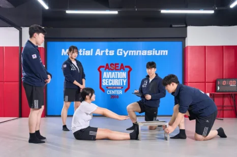 체육관 바닥에 앉은 두 사람과 주위 네 사람이 다리와 운동 도구를 살피고 있는 모습