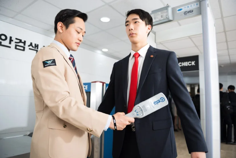 베이지색 제복 차림 사람이 정장 차림 사람의 몸 앞에 휴대용 검색기를 대고 있는 모습