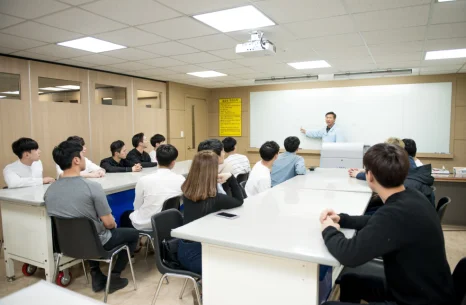 교실 책상에 여러 사람이 앉고 앞에 선 사람이 설명하고 있는 모습