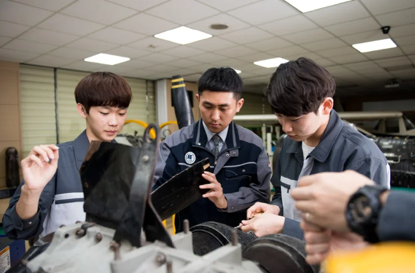 실습복 차림 여러 사람이 엔진 옆에서 도구를 보며 설명을 듣고 있는 모습