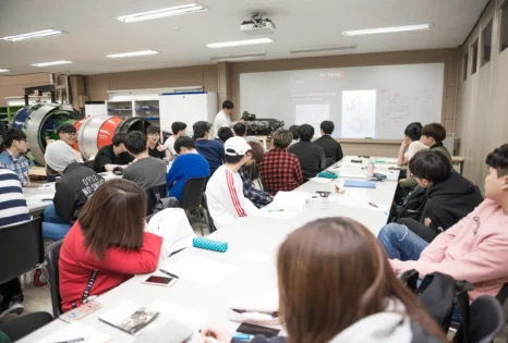 교실의 긴 책상에 여러 사람이 앉아 앞쪽 화면과 엔진을 보고 있는 모습