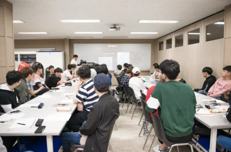 교실의 긴 책상에 여러 사람이 앉아 앞에 선 사람의 설명을 듣고 있는 모습