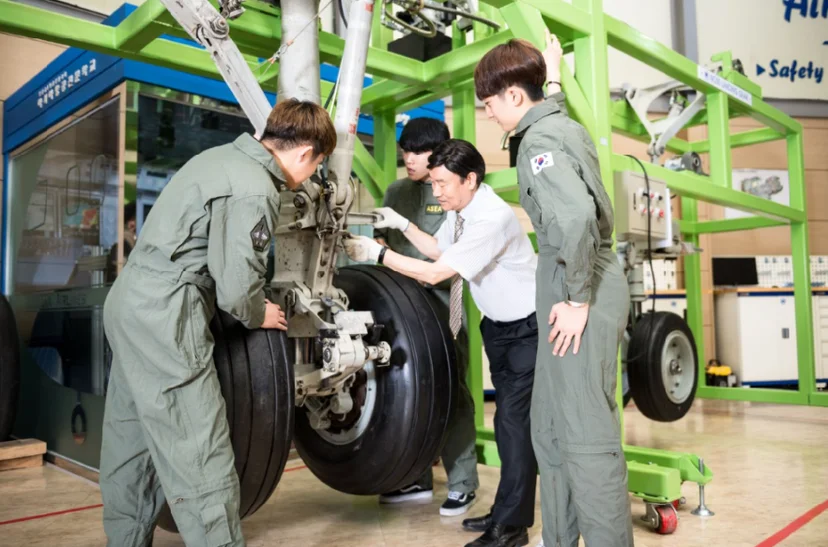 초록색 구조물 안 항공기 바퀴 옆에서 세 사람이 설명을 듣고 있는 모습