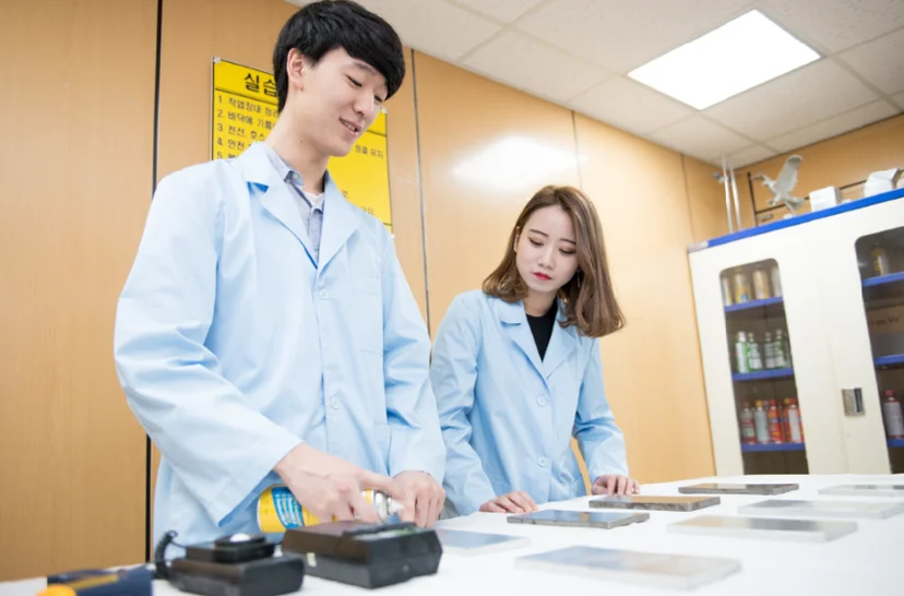 실습복 차림 두 사람이 작업대에서 금속 부품과 도구를 사용하고 있는 모습