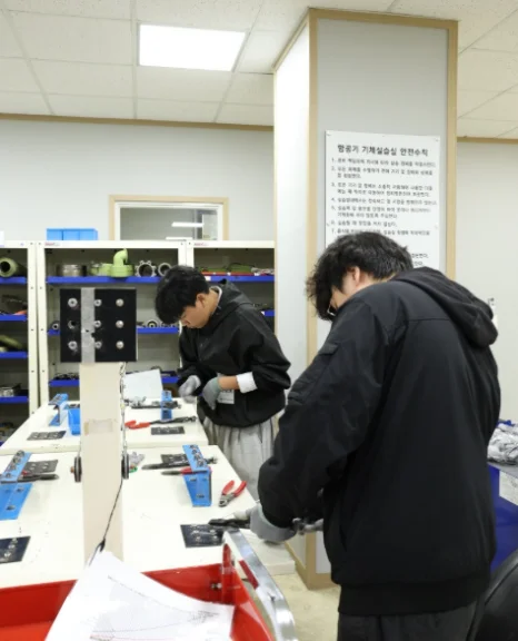 검은 옷 차림 두 사람이 작업대 위의 작은 부품과 도구를 사용하고 있는 모습