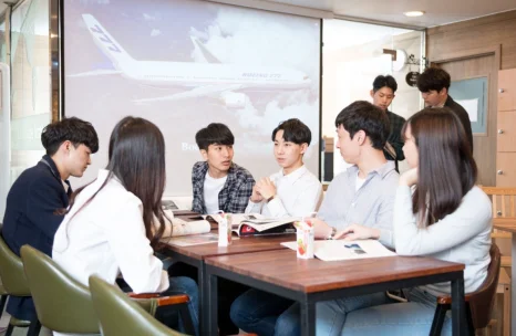 여러 사람이 책상에 둘러앉아 손으로 설명하고 뒤쪽 화면에 영상이 보임