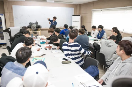 교실 책상에 여러 사람이 앉아 앞에 선 사람이 가리키는 장치와 화면을 보고 있는 모습
