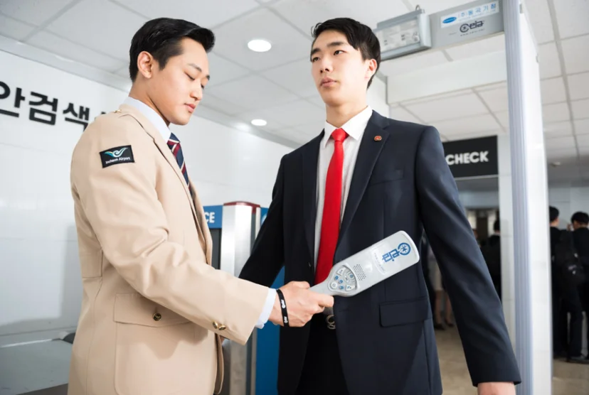 제복 차림 사람이 정장 차림 사람의 몸 앞에 손잡이 달린 검색 장비를 대고 있는 모습
