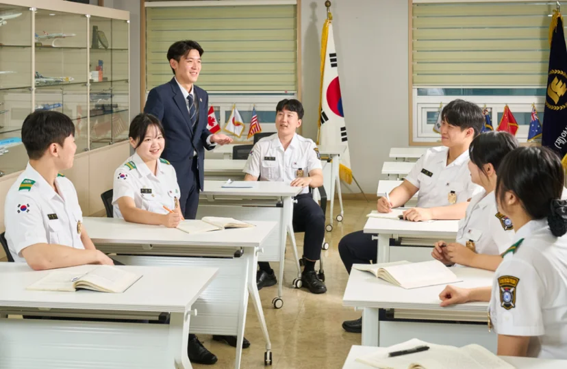 제복 차림 여러 사람이 교실에 앉고 정장 차림 사람이 서서 설명하고 있는 모습