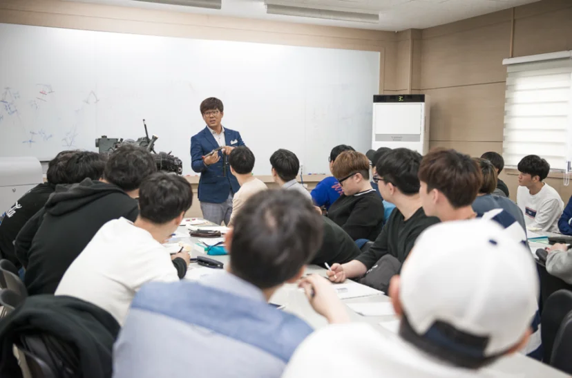 여러 사람이 교실의 큰 책상에 둘러앉아 앞에 선 사람의 설명을 듣고 있는 모습