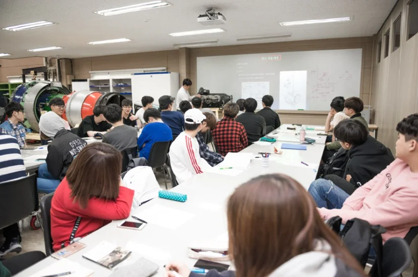 긴 책상에 앉은 여러 사람이 실습실 앞쪽 설명을 듣고 있는 모습
