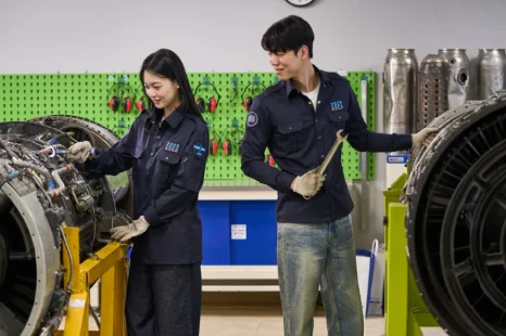 실습복 차림 두 사람이 엔진 옆에서 손과 도구로 부품을 살피고 있는 모습