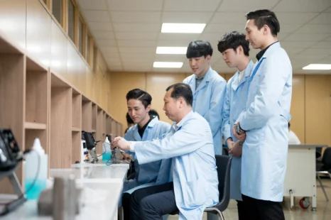 흰 실습복 차림 여러 사람이 앉은 사람의 작업을 지켜보고 있는 모습