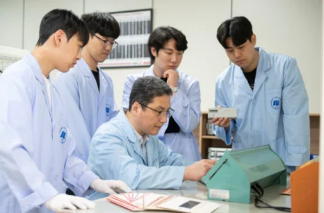 흰 실습복 차림 여러 사람이 앉은 사람과 책상 위 부품을 함께 살피고 있는 모습