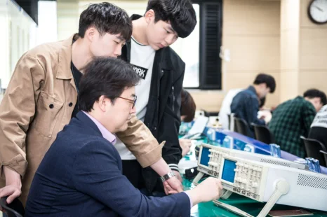 정장 차림으로 앉은 사람이 흰 전자 장비를 조작하고 여러 사람이 지켜보고 있는 모습