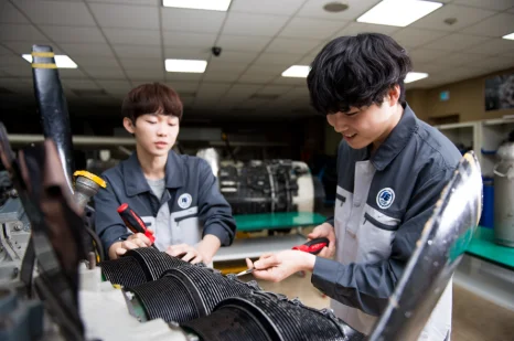 실습복 차림 두 사람이 검은 부품에 도구를 대고 있는 모습