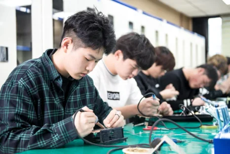 작업대에 앉은 여러 사람이 작은 전기 부품에 도구를 대고 있는 모습